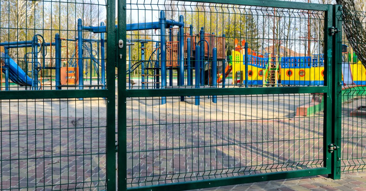 A closed metal gate outside of a children's playground. There are wide gaps between the wires in the fence.