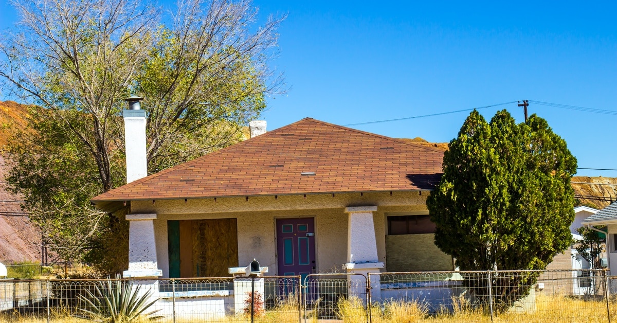 A home has its windows boarded up and the door shut with a padlock. There is a chain link fence surrounding the house.