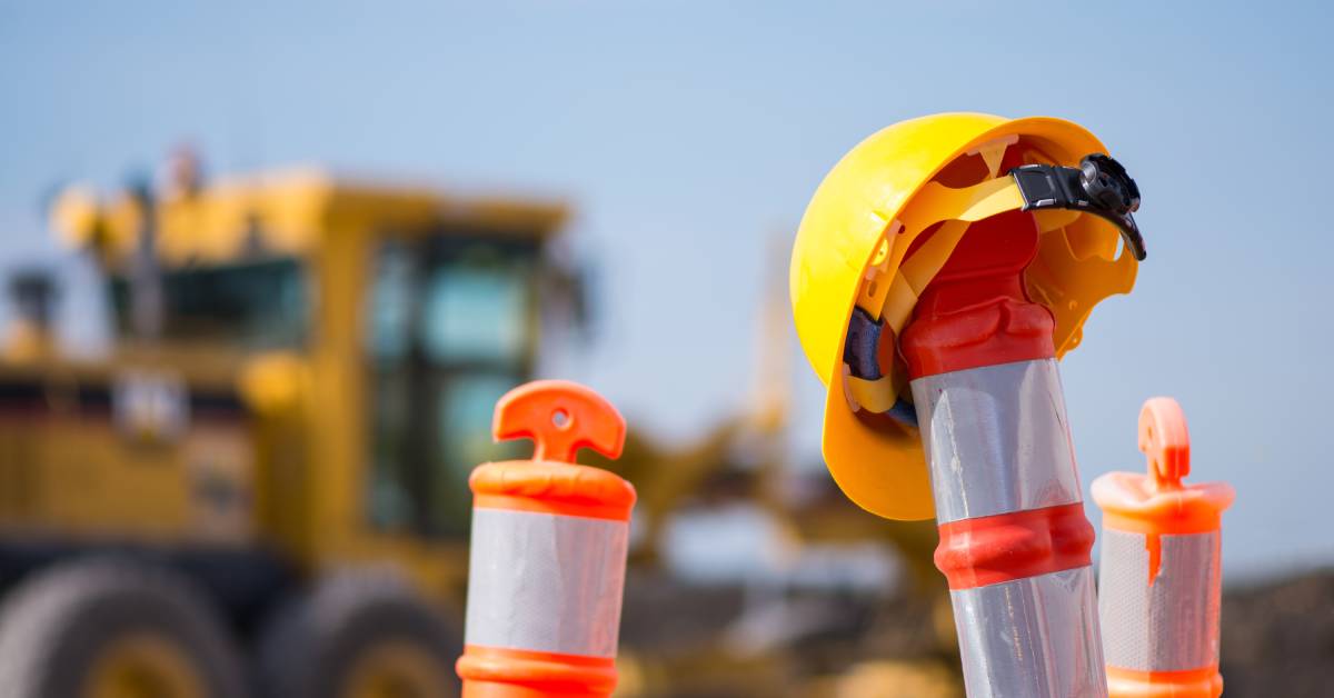 A hard hat on the top of a construction cone with others behind it. Further back is larger construction equipment.