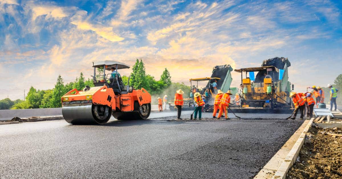 Construction crew working on a highway road with large equipment moving around. The crew is laying asphalt.