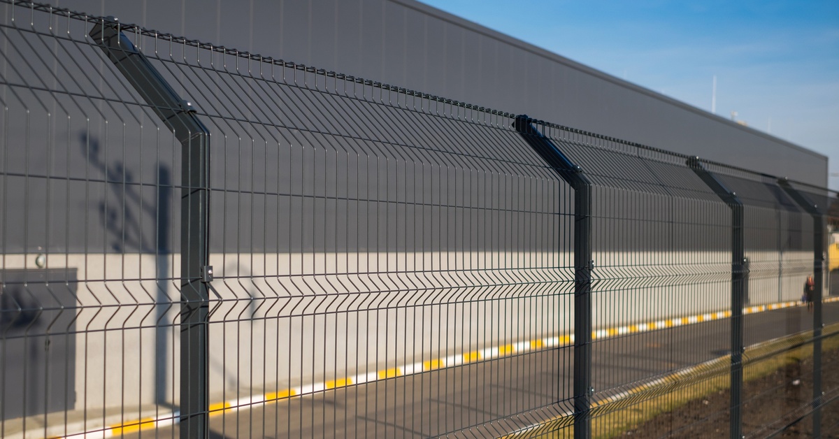 A steel fence with a curved top to keep intruders away. There is a road and building behind the fence.