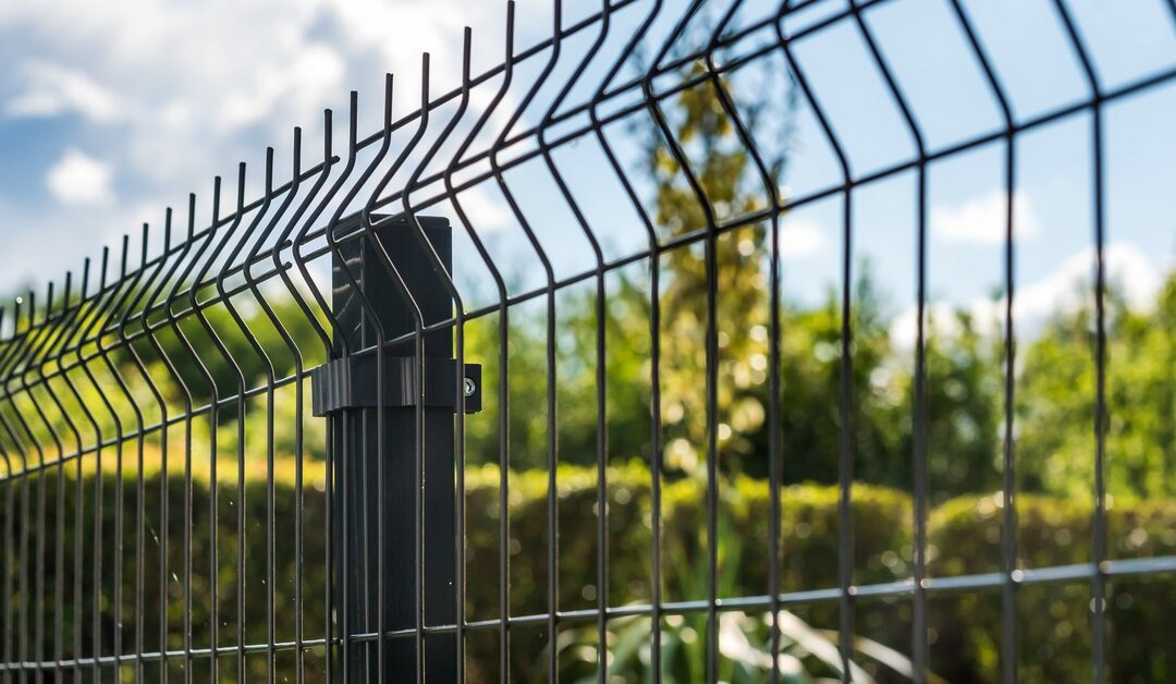 A black fence with grating wire running across it. There are blurred-out trees and bushes in the background.