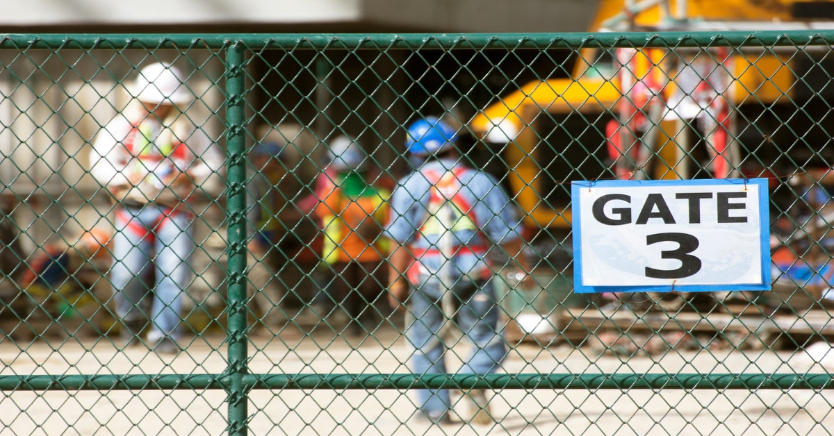A fence with a "gate three" sign attached to it. Behind the fence are construction workers and their equipment.