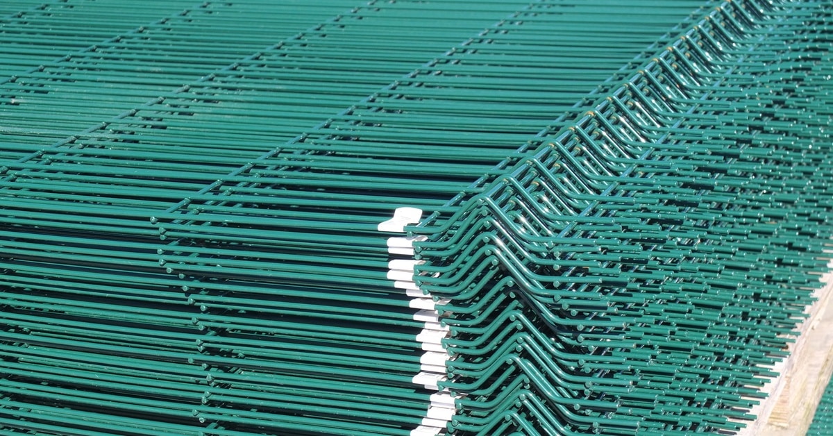 A stack of green, curvy welded fencing panels. These panels are sitting on a pallet inside a warehouse.