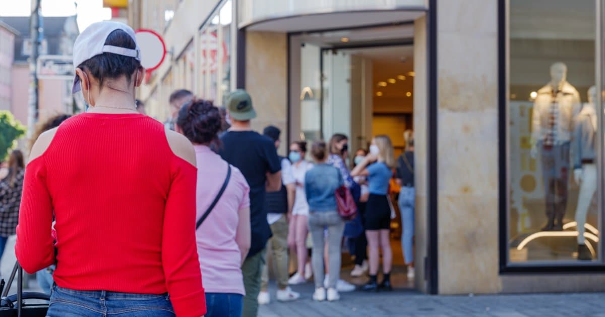 People standing in a line outside of a large store waiting to enter. One person in the crowd has a bright red shirt on.