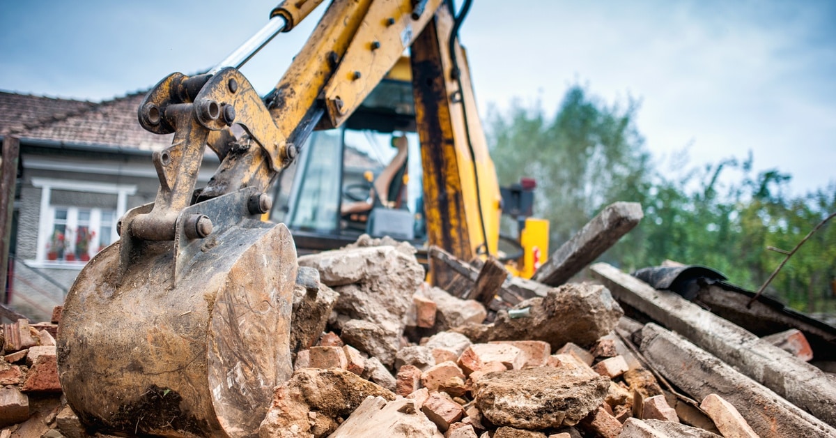An excavator picks up pieces of a demolished building that are in a pile. A house is visible behind the excavator.