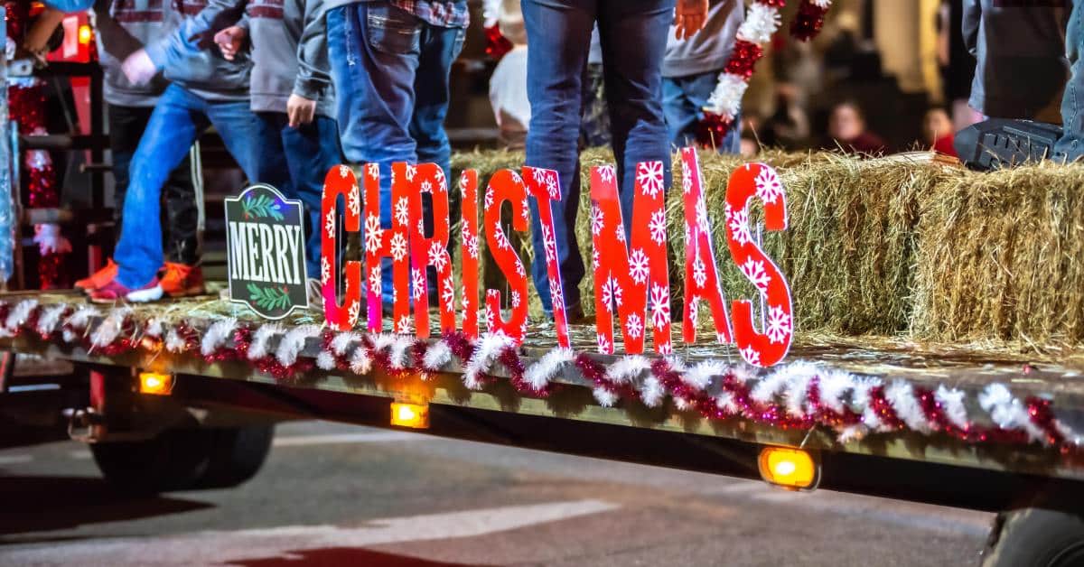 A parade float with a Merry Christmas sign on it and hay bales. People are also standing on the float.