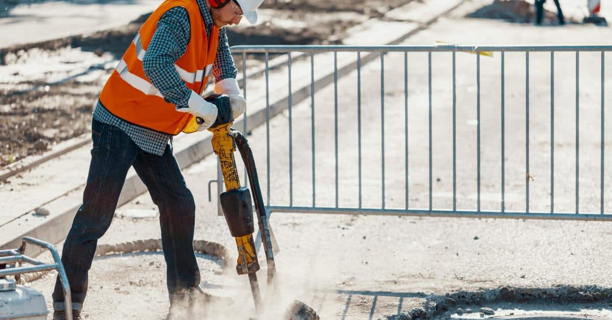 A person on a construction site using a drill on a street with a metal fence behind him. He is wearing safety gear.