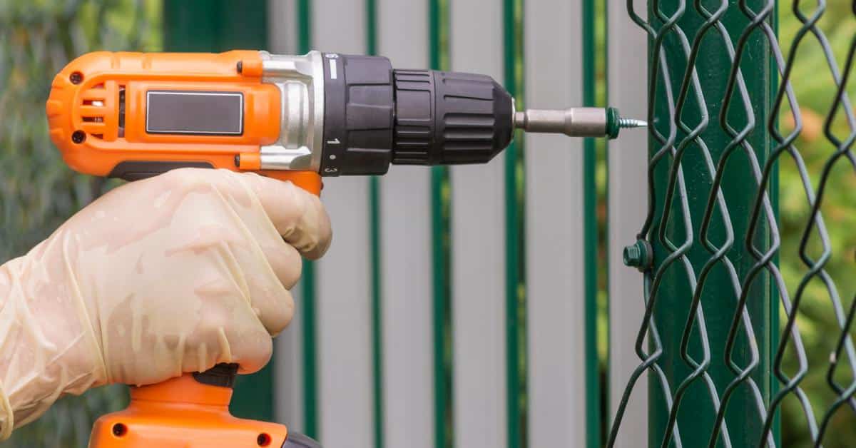 A person using an electric drill to push a nail into boards on a metal fence. Next to the boards is chain-link fencing.