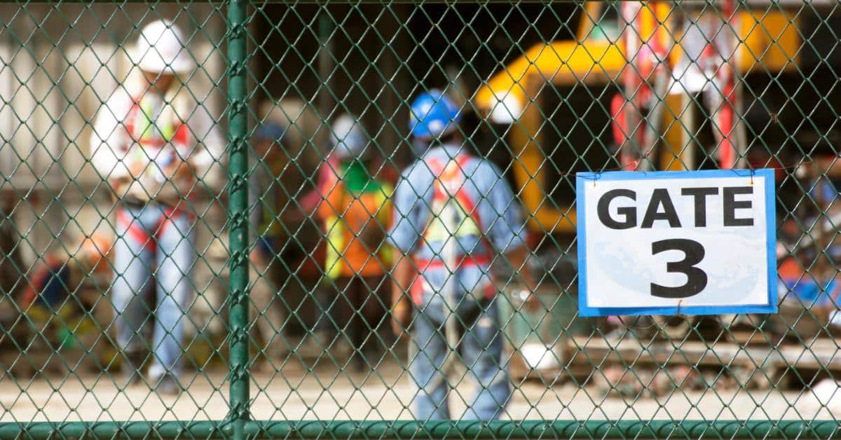 Workers on a construction site, blocked by a chain-link fence. There is a sign on the fence that says gate 3.