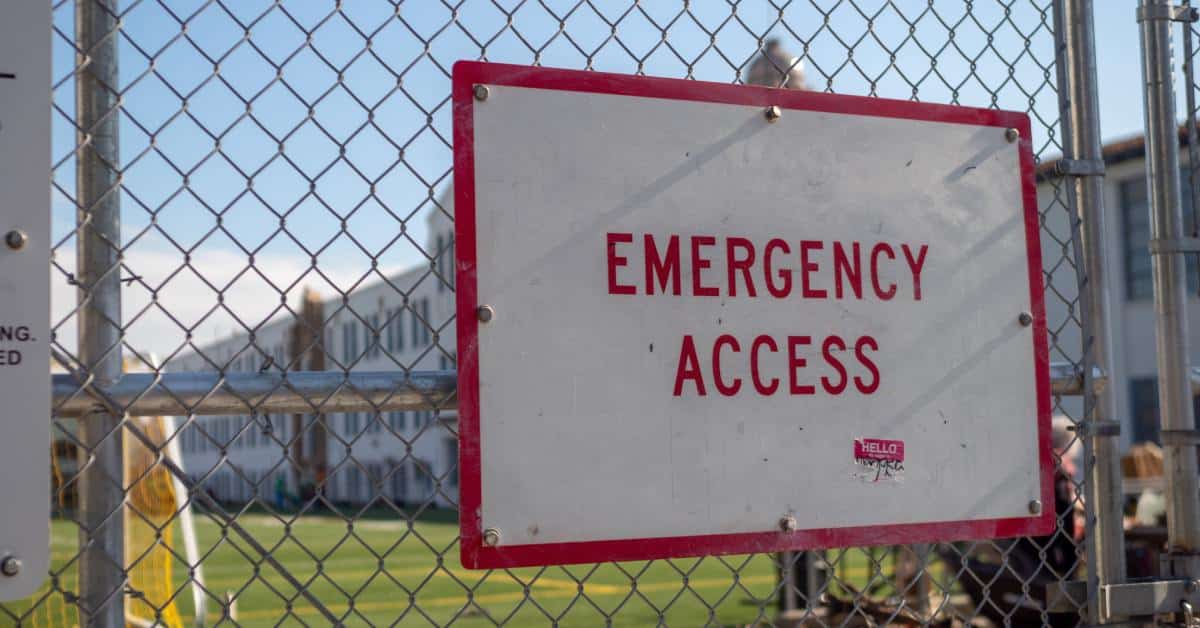 A chain-link fence preventing unauthorized entrance to a school field. The fence has a sign that says emergency access.